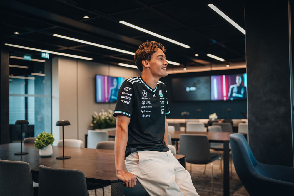 George Russell, jezdec týmu Mercedes-AMG PETRONAS Formula One Team, neformálně sedí na stole v moderní a stylové týmové zóně nebo kanceláři. Usmívá se a má na sobě oficiální týmovou košili s viditelnými logy sponzorů, jako jsou Einhell, IWC a CrowdStrike. Na obrazovkách v pozadí je vidět jeho fotografie.