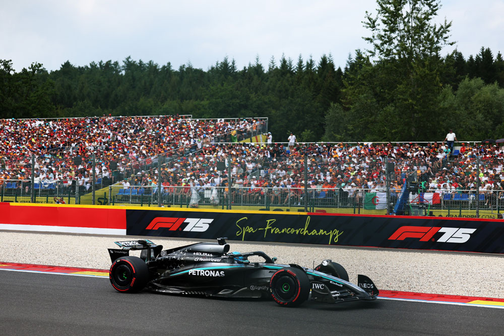 Vůz Mercedes-AMG PETRONAS Formula One Team jede po okruhu Spa-Francorchamps. V pozadí jsou plné tribuny fanoušků mávajících mezinárodními vlajkami. Logo Spa-Francorchamps je viditelné na bariéře.