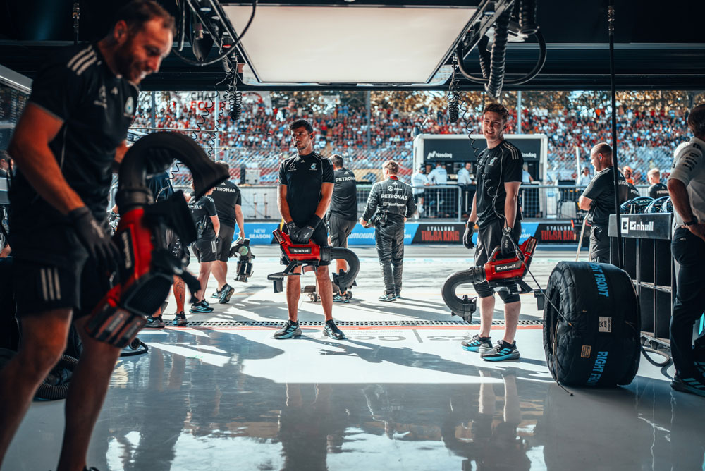 Mechanici týmu Mercedes-AMG PETRONAS Formula One Team se v garáži připravují s bezdrátovými foukači Einhell před zastávkou v boxech. V pozadí jsou tribuny plné fanoušků.