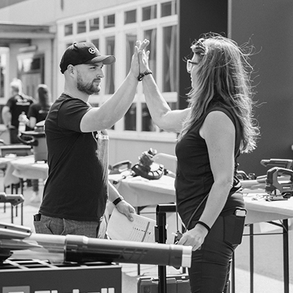 Muž a žena si dávají high five na venkovní akci Einhell s vystaveným nářadím, zobrazeno na černobílé fotografii.
