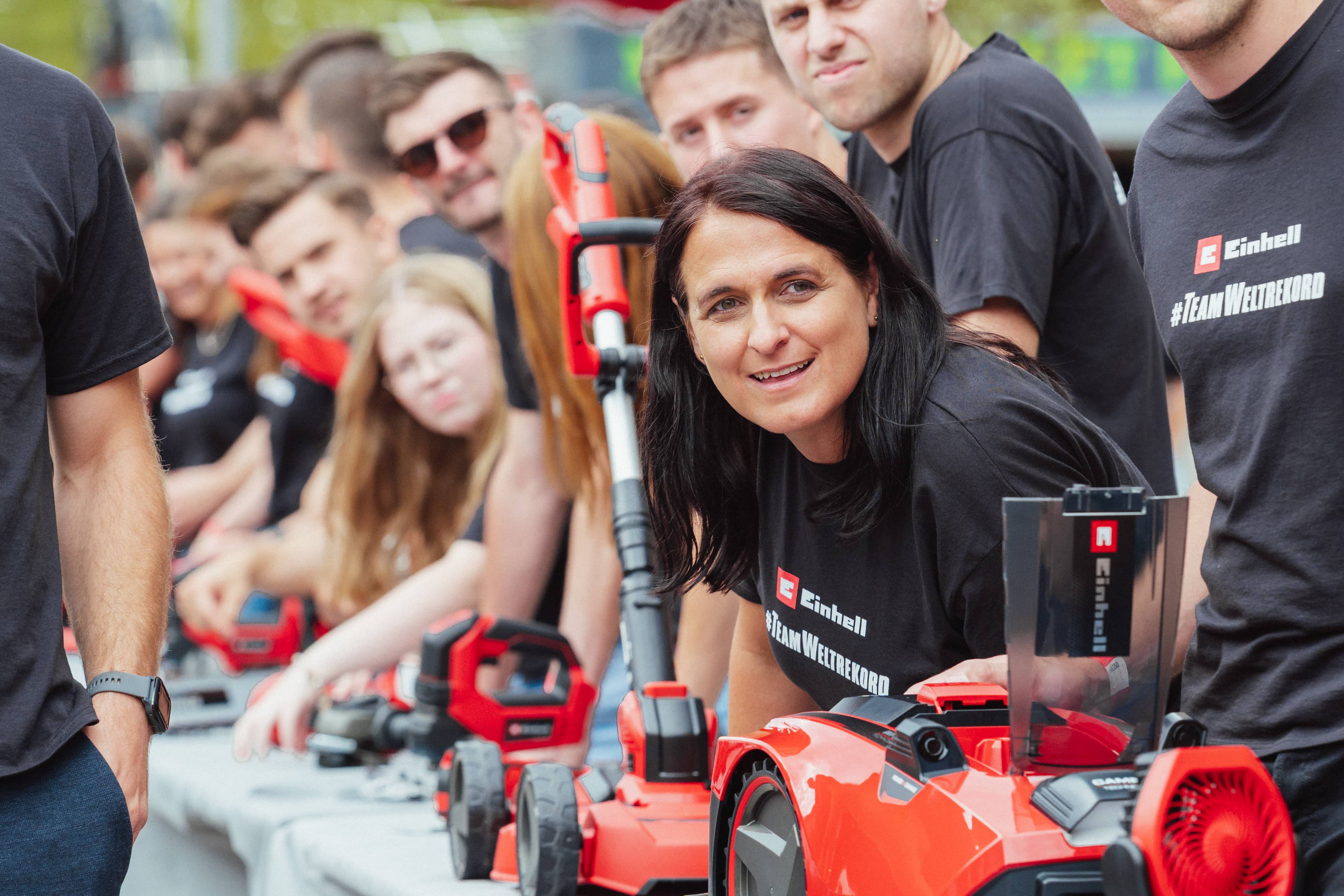 Žena se naklání dopředu a usmívá se do kamery, obklopena dalšími zaměstnanci Einhellu a zařízeními.