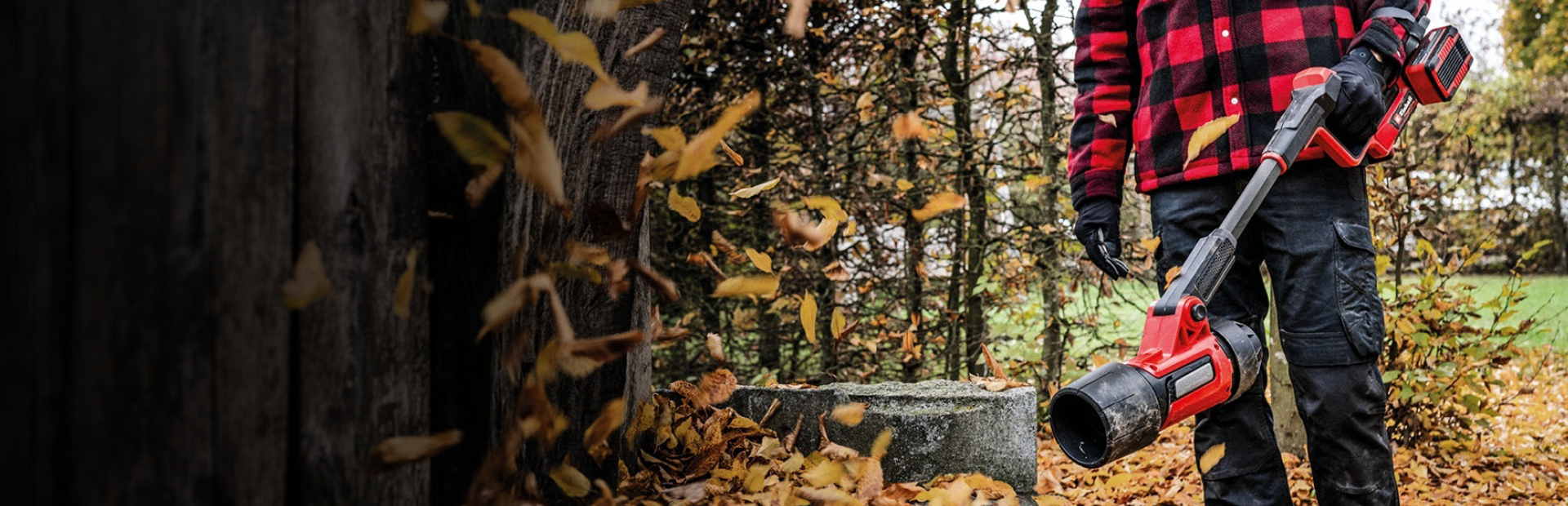 Osoba v čiernych nohaviciach, rukaviciach a červeno-čiernej károvanej bunde stojí vonku a drží červený akumulátorový fukár na lístie od Einhellu. Na zemi leží opadané jesenné lístie a v pozadí sú vidieť stromy.